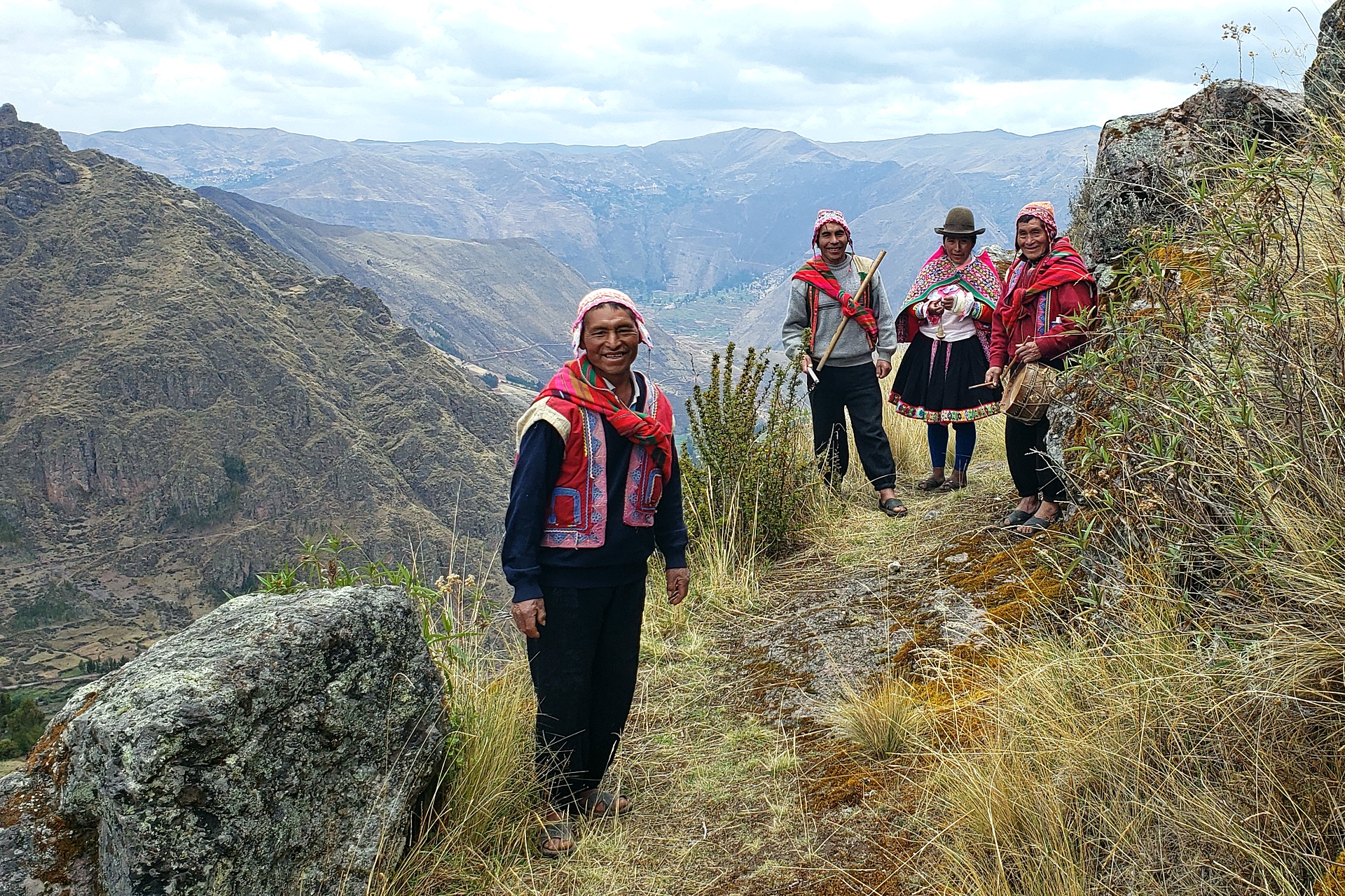 Four indigenous Peruvians in traditional attire shown smiling and playing instruments above Peru's Sacred Valley