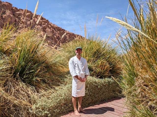 A man in a bathrobe surrounded by tall grasses and red rocks at Puritama Hot Springs