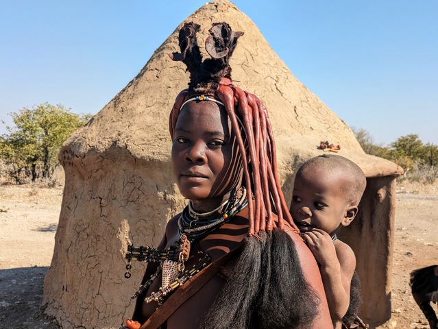 A Himba woman carrying her baby on her back with a mud hut behind her, as encountered on a luxury safari in Namibia