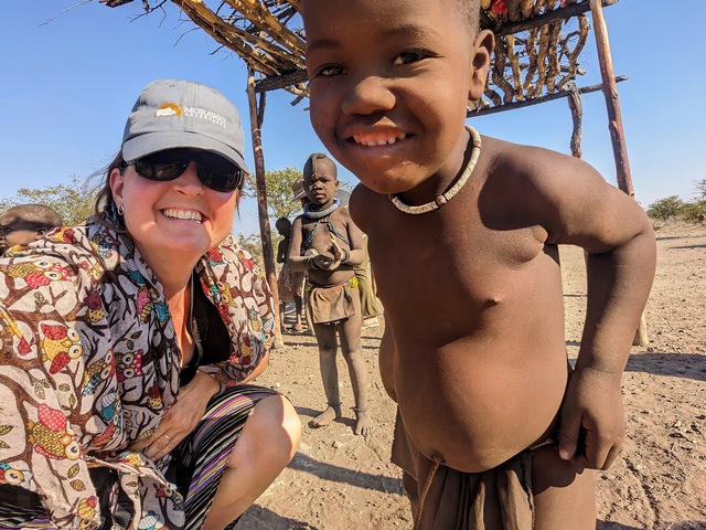 A safari guest and Himba child smiling at the camera as another Himba child looks on with curiosity from behind