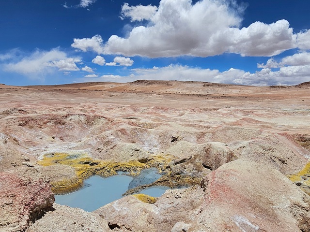 Colorful pools of mud and mineral deposits against a red rocky background and blue sky with puffy clouds, el geyser sol de manana