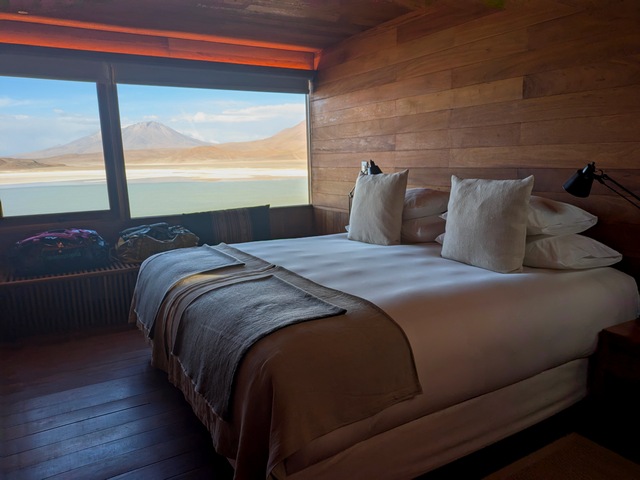 A freshly made bed inside a wood-paneled room with huge picture windows showcasing an emerald lake and volcano at Explora Ramaditas Lodge