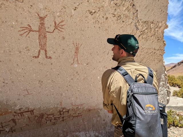 A hiker stands next to ancient pictographs showing human and animal figures in the valley of the yardangs on Explora's Travesia