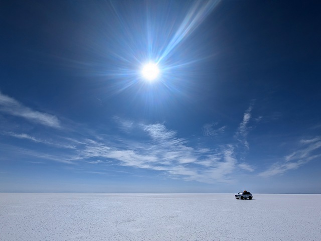 A single vehicle looks tiny compared to the endless expanse of flat and white that surround it under a sunny blue sky at the uyuni salt flats