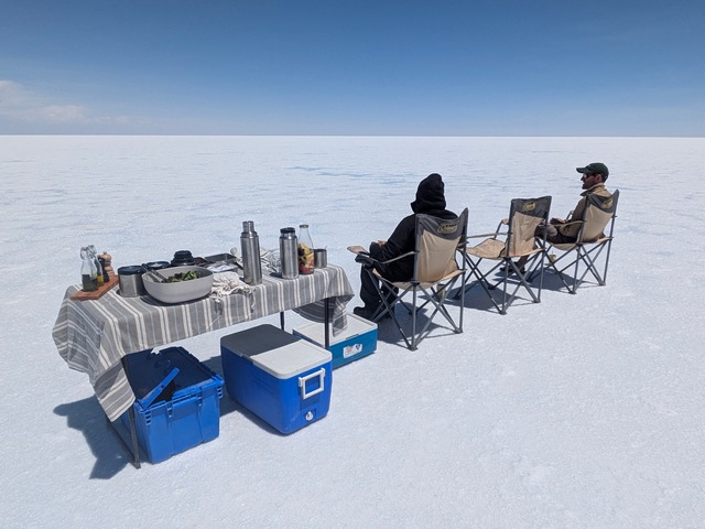 Two people enjoying a picnic lunch while seated in folding chairs next to a table on the vast, endless Uyuni Salt Flats