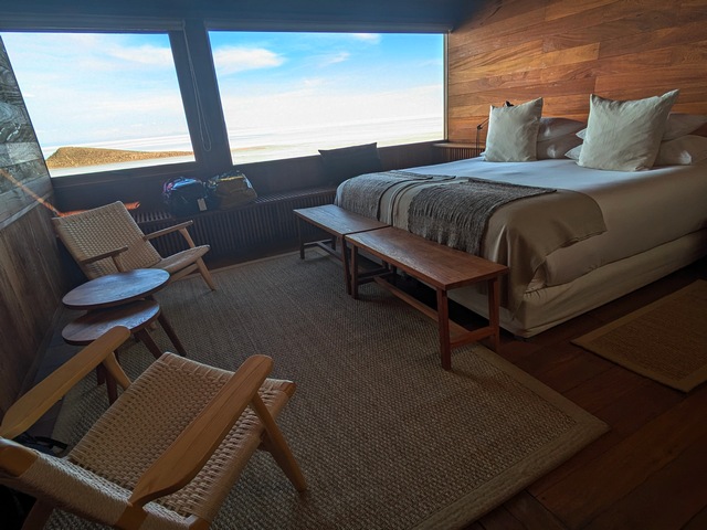 Guest bedroom inside Explora Uyuni Lodge, with a bed and two chairs next to a large picture window with a view of the Uyuni Salt Flats outside