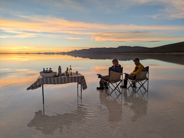 A couple sitting in chairs next to a table, enjoying sundowner drinks against a colorful and reflective view of the sky and landscape at sunset in the Uyuni Salt Flats