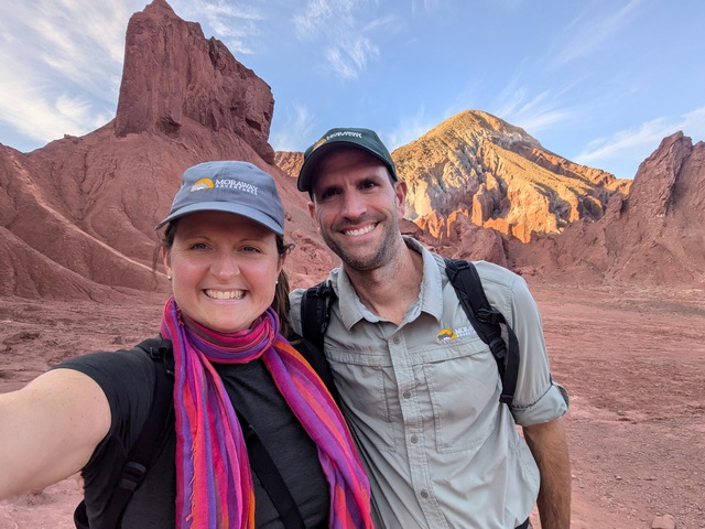 a selfie of two hikers with the Atacama Desert's rocky red landscape of the Rainbow Valley behind them