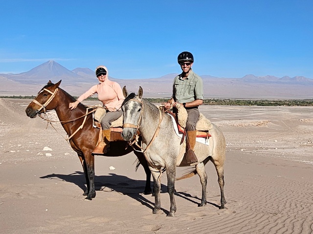 two people on a horseback ride in the Salt Valley of San Pedro de Atacama, with sand dunes and Volcan Licancabur behind them