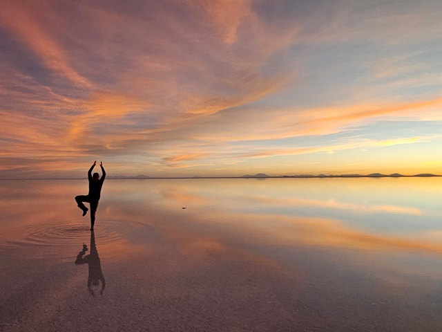 A person in a yoga pose at sunset on the uyuni salt flats, with the person and clouds reflecting in the water