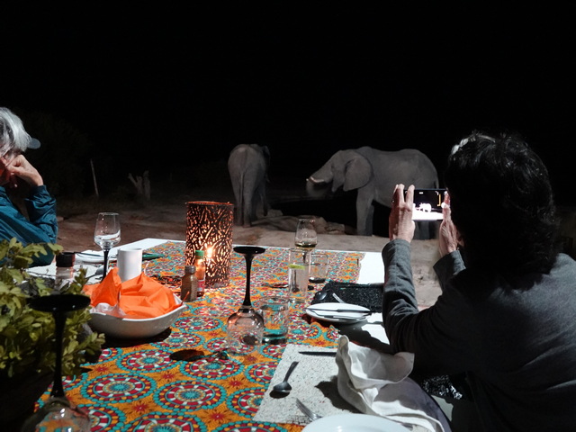 Two guests photograph elephants at the waterhole during dinner at Nehimba Camp, a luxury safari camp in Hwange National Park Zimbabwe