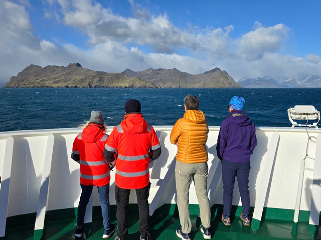 four cruise ship passengers stand on the bow of the ship and look out at the beautiful, jagged, mountainous landscapes of South Georgia Island