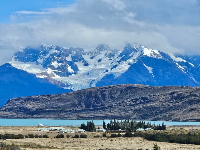 far away view of the buildings of estancia cristina on the shores of the turquoise Lago Argentina, with large glaciated mountains in the background