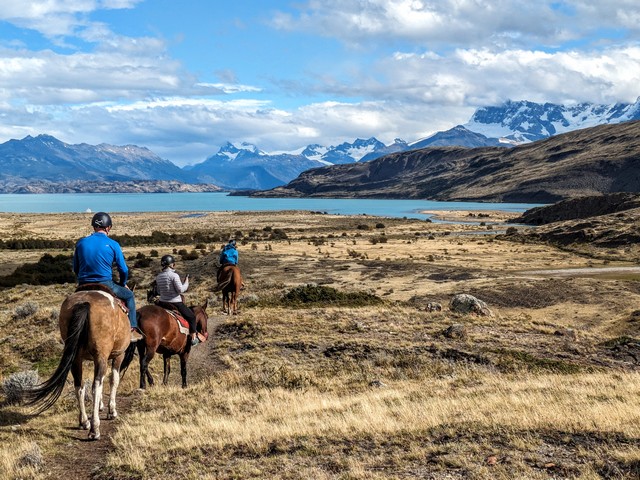 three horseback riders head across the Patagonian steppe, toward the teal colors of lago argentina and the mountains beyond at estancia cristina