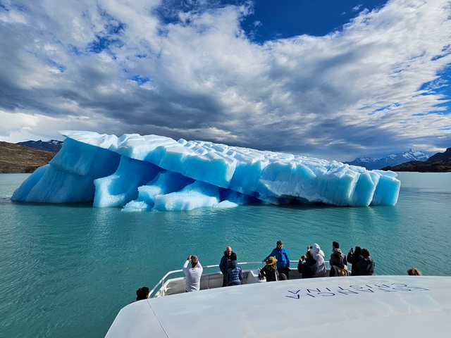 a vibrant, blue slanted iceberg floats in the aquamarine colored waters of lago argentina as several tourists look on from a catamaran