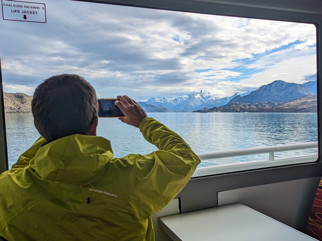 a man taking photos out a big picture window of a catamaran on the calm waters of lago argentina, surrounded by towering mountains