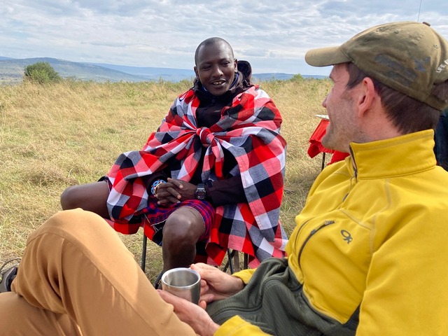 a maasai warrior and a safari guest sit in picnic chairs talking with the plains of the Maasai Mara visible behind them