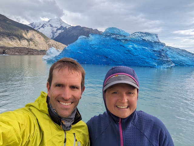 two smiling tourists on a catamaran in lago argentina, with a beautiful blue iceberg and towering glaciated peak behind them