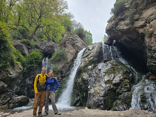 two hikers pose in front of a waterfall and lush trees in Morocco's High Atlas Mountains