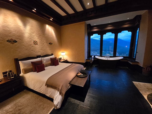 Guest room at Gangtey Lodge in Bhutan featuring warm wood interiors, a cozy bed, ambient lighting, and panoramic views of the Phobjikha Valley through a large window.