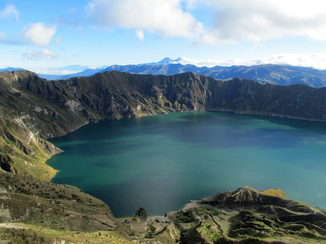 View from the rim of Quilotoa Crater in Ecuador, overlooking the turquoise crater lake under a blue sky with scattered white clouds, with the distant Illinizas peaks faintly visible beyond the rim.