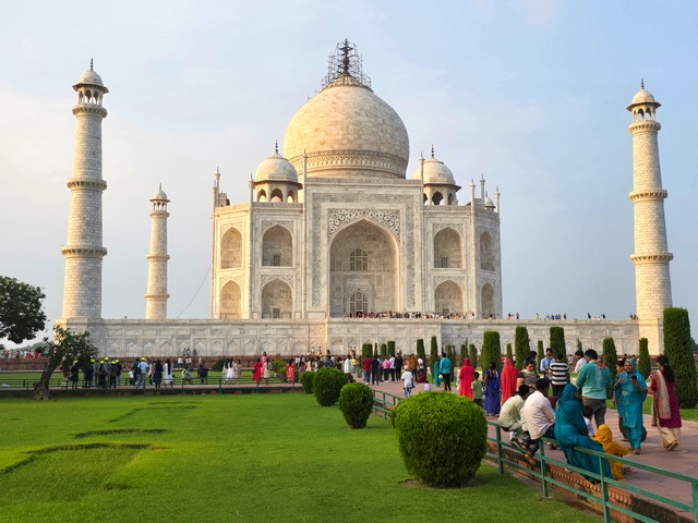 Wide view of the Taj Mahal in Agra, India, showing the full front façade and surrounding Mughal gardens with visitors walking along the pathways under soft evening light and a clear sky