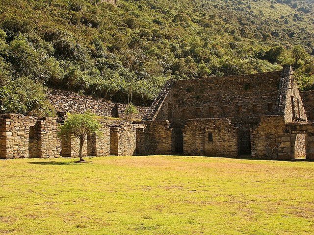Stone ruins at Choquequirao in Peru, featuring a well-preserved Inca building and adjacent stone walls overlooking a green courtyard, with lush vegetation covering the surrounding mountainside.