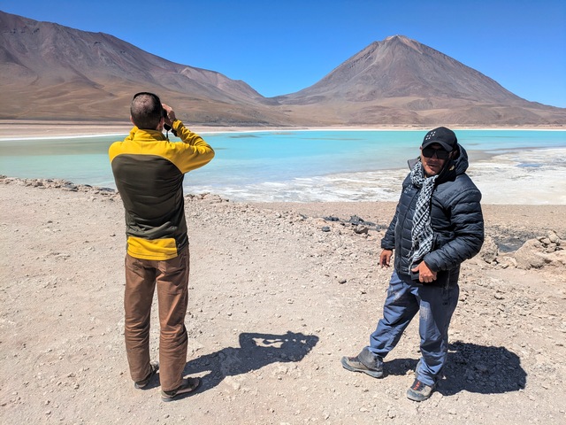Laguna Verde in Bolivia on the Atacama to Uyuni Explora Travesía route, showing turquoise-green lake waters with a stark brown volcano in the background as a traveler looks through binoculars beside his guide