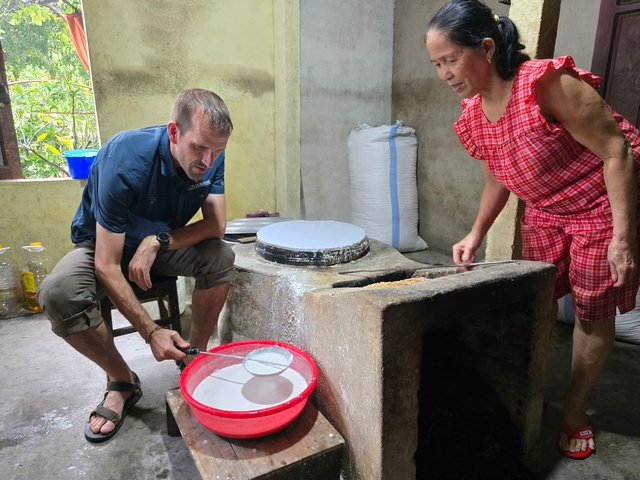 Inside a Vietnamese cooking workshop, an American man learns to make traditional rice paper by ladling rice batter onto a hot griddle while an older Vietnamese woman demonstrates and supervises with a smile.