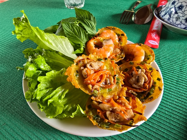 Close-up of a vibrant meal in the Mekong Delta, Vietnam featuring bánh khọt topped with shrimp and vegetables alongside a large serving of fresh greens including lettuce and basil.
