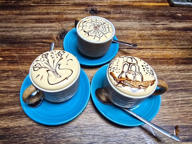 Three blue mugs of Vietnamese egg coffee with creamy foam and detailed latte art made with chocolate syrup, including designs of a ship, spider web, and flowers, served on matching saucers in Hanoi.