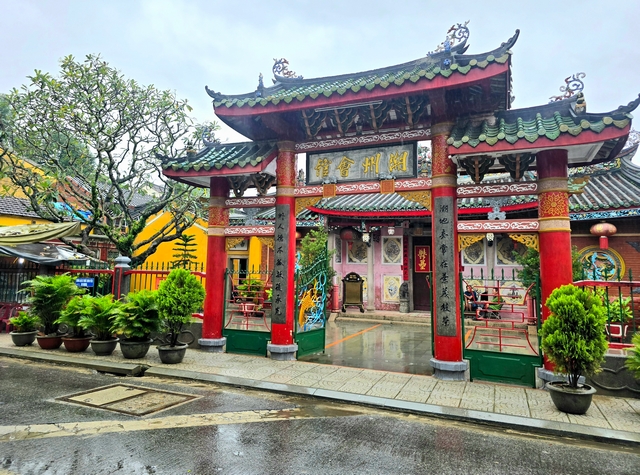 Street view of Hoi An’s Chaozhou Hall in Vietnam on a rainy day, showing the colorful entrance gate with red, pink, and yellow details and green potted plants, resembling a traditional pagoda.