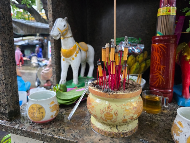 Close-up of offerings inside a spirit house hanging from a tree near Hoi An market, Vietnam, including a golden ceramic incense pot with incense sticks and cigarettes, a white horse statue with a gold saddle, and various ceremonial vessels