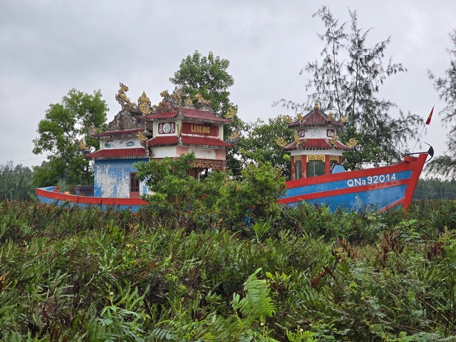 Boat temple in Duy Vinh near Hoi An, Vietnam (Bon Van Duy Vinh), featuring a blue and white ship-like temple with red accents surrounded by lush tropical vegetation, trees, and ferns.