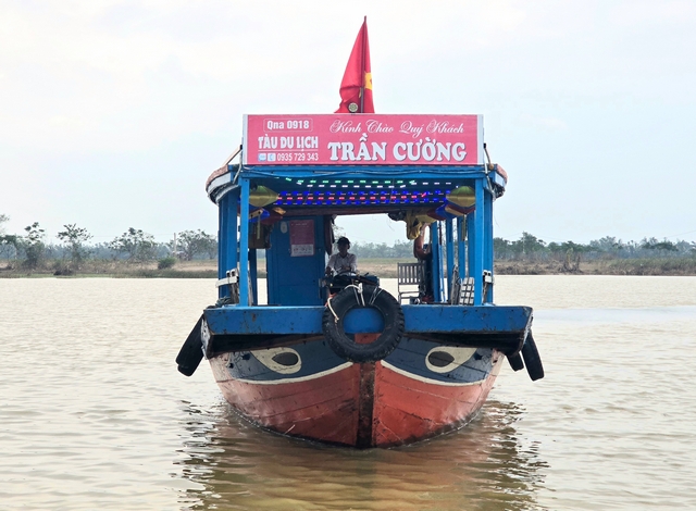 Head-on view of a traditional wooden tourist boat on the Thu Bon River near Hoi An, Vietnam, featuring a red hull, blue accents, and painted eyes on the bow, floating on brown river water.