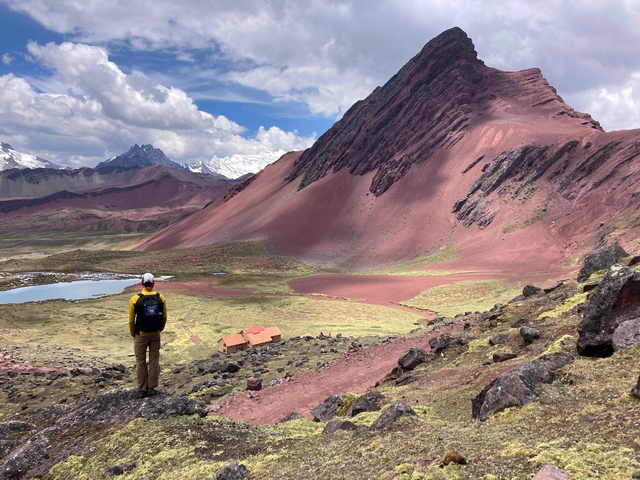 A hiker gazes out at stunning redrock mountain formations near Peru's sacred Apu Ausangate, with the community-run Huampococha Lodge visible in the valley bottom below.