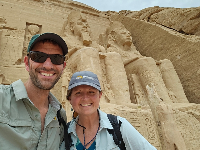 A smiling couple standing next to two giant stone statues of Ramesses II at the entrance to the Great Temple at Abu Simbel, Egypt.
