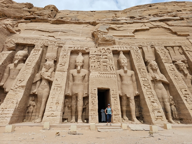A traveler standing inside the door at the main entrance of the Small Temple of Hathor and Nefertari at Abu Simbel, flanked by three giant carved statues of Nefertari and Ramesses II on either side of the door.