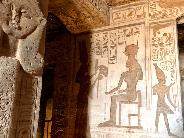 A glimpse inside the Small Temple of Hathor at Abu Simbel, Egypt, showing a carved stone head of the goddess Hathor on a pillar, and a stone relief carving showing Nefertari making an offering to a seated Hathor.