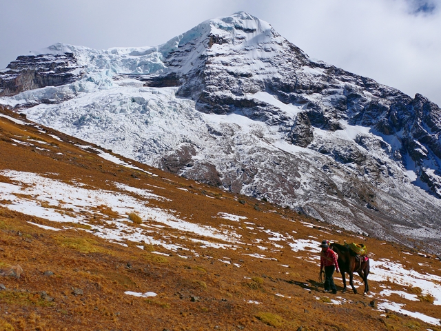 An Andean man with his horse stand in front of the towering, rocky, glaciated face of Peru's Ausangate mountain.