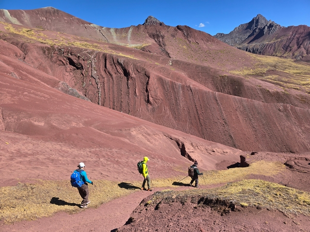 Three hikers walk down a smooth trail, surrounded by a mountainous landscape of deep red rocky peaks near Huampococha Lodge and the Red Valley, Peru.