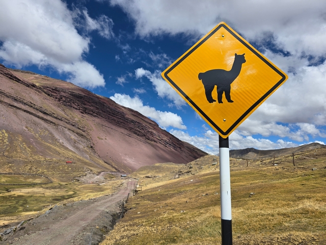 A yellow road sign indicating alpaca crossing, with cloudy blue skies and red mountains behind it.