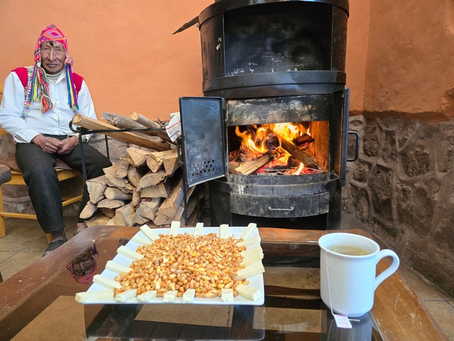 An Andean man sits next to a wood-burning stove, with a drink and corn snack on the table next to him.