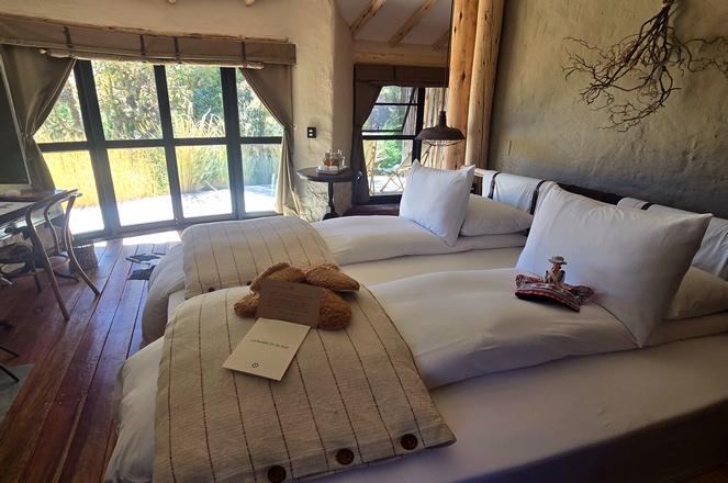 Bedroom inside a room at PUQIO by Andean in Colca Canyon Peru, showing a beautifully set queen bed with alpaca comforter, welcome gifts, and greenery outside the wooden-framed window.