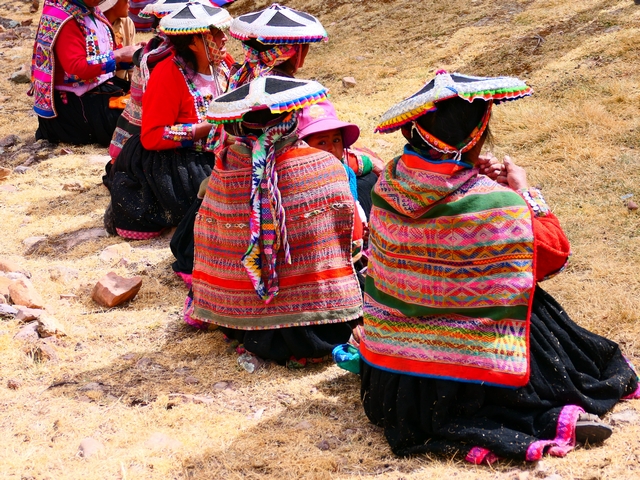 Six women in traditional Andean dress sit on the ground while spinning wool, wearing beautiful hand-woven garments characteristic of the Pitumarca region of Peru.
