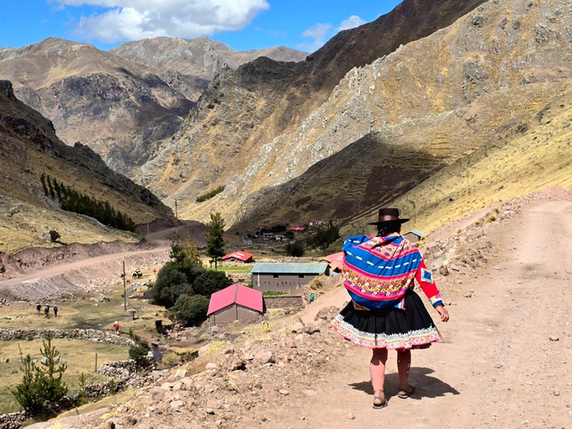 A Quechua woman walks down a dirt road toward a small village, wearing a black skirt, black hat, and carrying a woven bundle on her back.