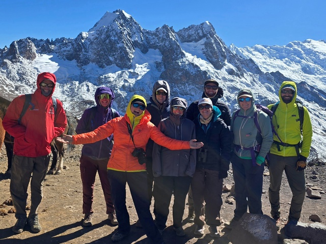 A group of nine trekkers bundled up in jackets on a sunny but windy day in Peru, with huge glaciated peaks and Ausangate behind them.