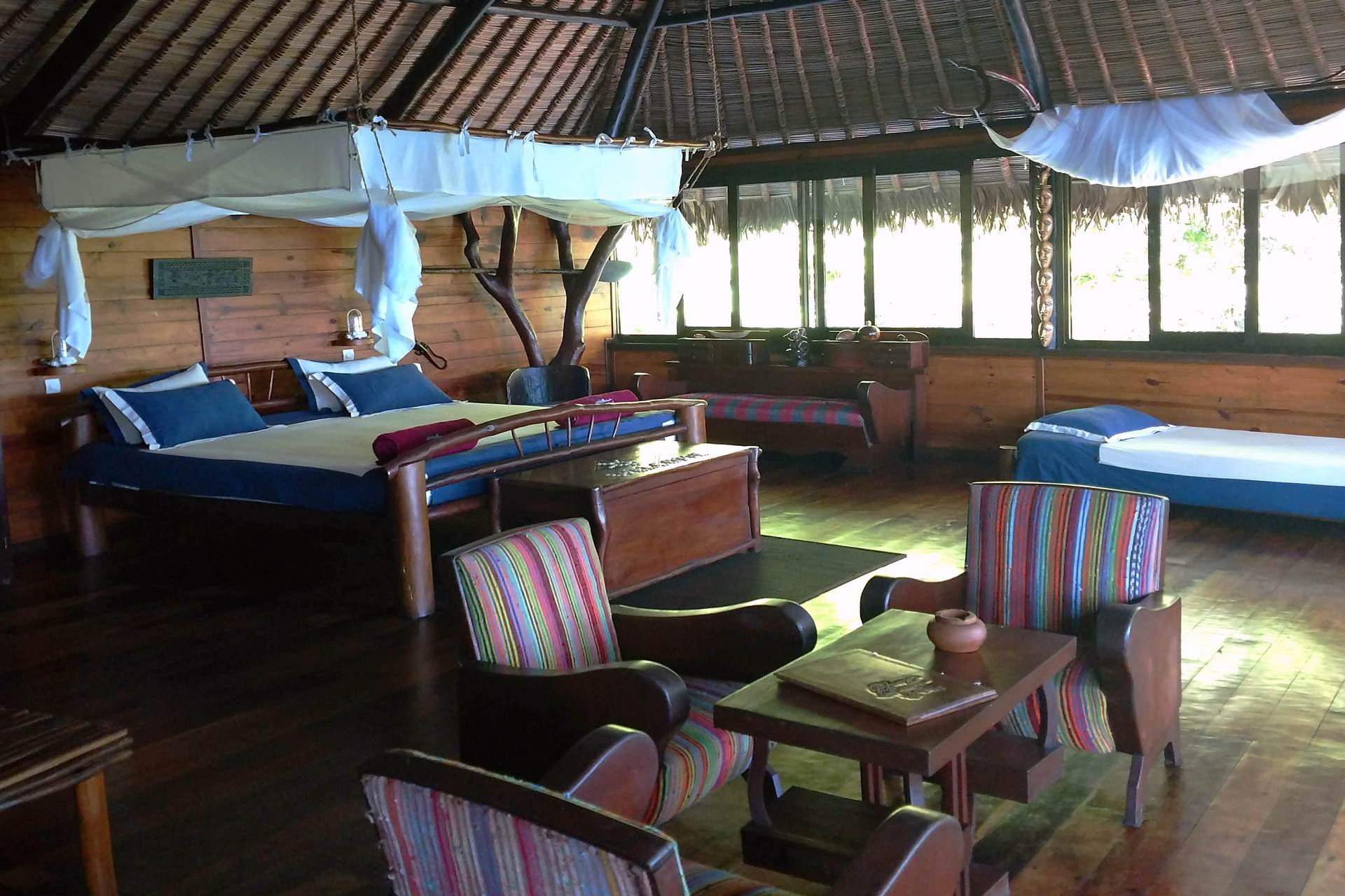 A guest room at Madagascar's Manafiafy Forest Lodge, showing wood-paneled floors and walls, a canopy bed with mosquito netting, and colorfuly upholstered furniture.